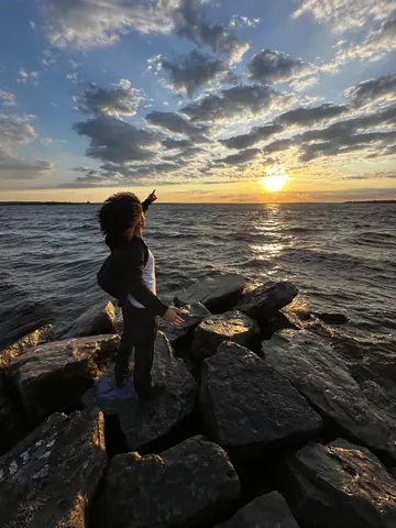 Taha standing on shoreline rocks at sunset, pointing up at the sky over the water.